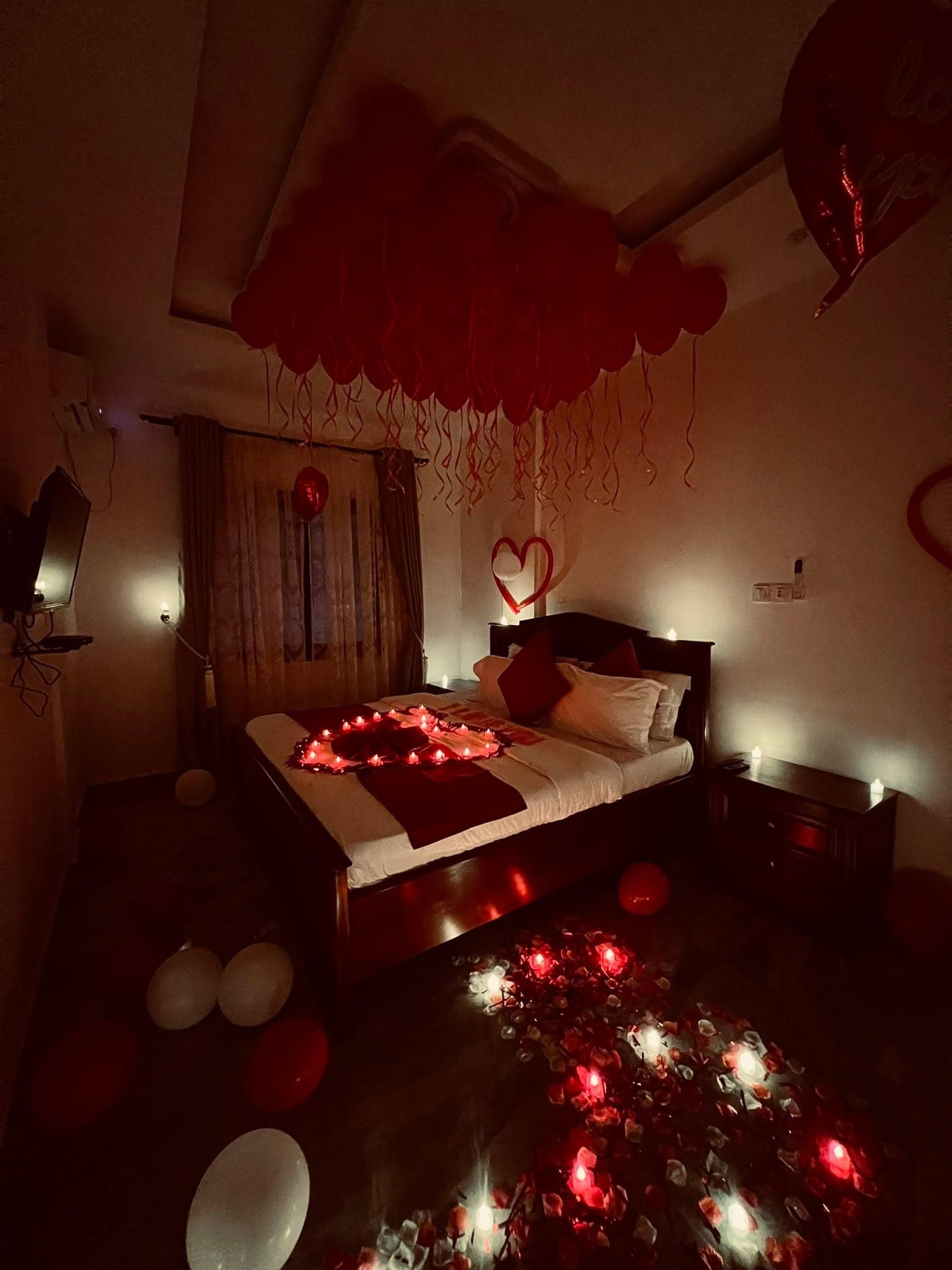Chambre de l’appartement dressée pour la Saint-Valentin — lit en bois sombre, voûte de ballons rouges, téléviseur mural et fenêtre voilée