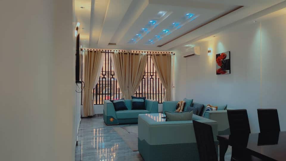 Salon de l’appartement A10 — canapés et fauteuils en velours bleu-vert, coussins baroques dorés et bleu nuit, sol en marbre, faux-plafond à gorge lumineuse bleue, climatiseur mural, doubles rideaux et coin repas au fond