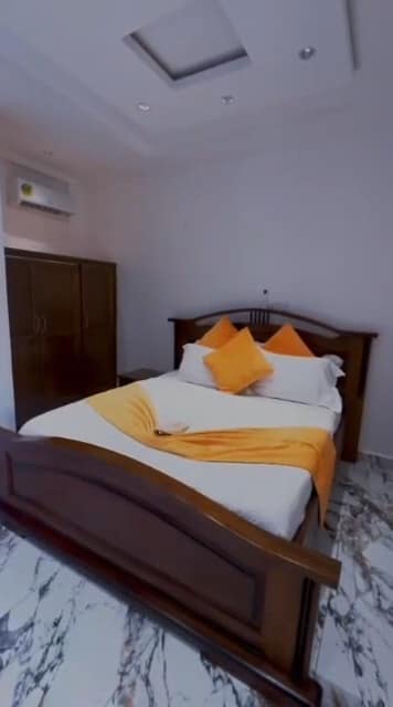 Chambre de l’appartement A10 — grand lit en bois sombre, linge blanc et orange, armoire, climatiseur mural et sol en marbre
