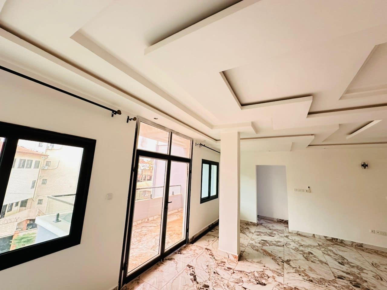 Salon vide de l’appartement A40 — sol en marbre veiné rose et or, faux-plafond à redans avec bandeaux lumineux, poteau central, baie vitrée à châssis noir ouvrant sur le balcon et tringles à rideaux posées