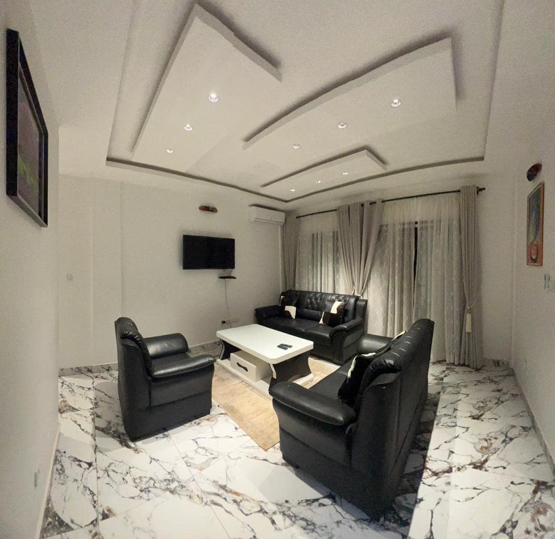 Salon du B35 — canapé et fauteuils en cuir noir capitonné, table basse blanche, téléviseur mural, climatiseur, faux-plafond à caissons et spots encastrés, sol en marbre blanc veiné