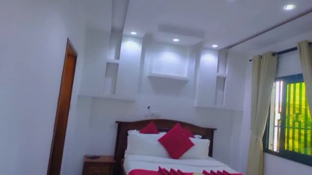 Chambre de l’appartement A10 — grand lit en bois verni, linge blanc et coussins framboise, niches et spots au plafond, fenêtre à rideaux crème et sol en marbre