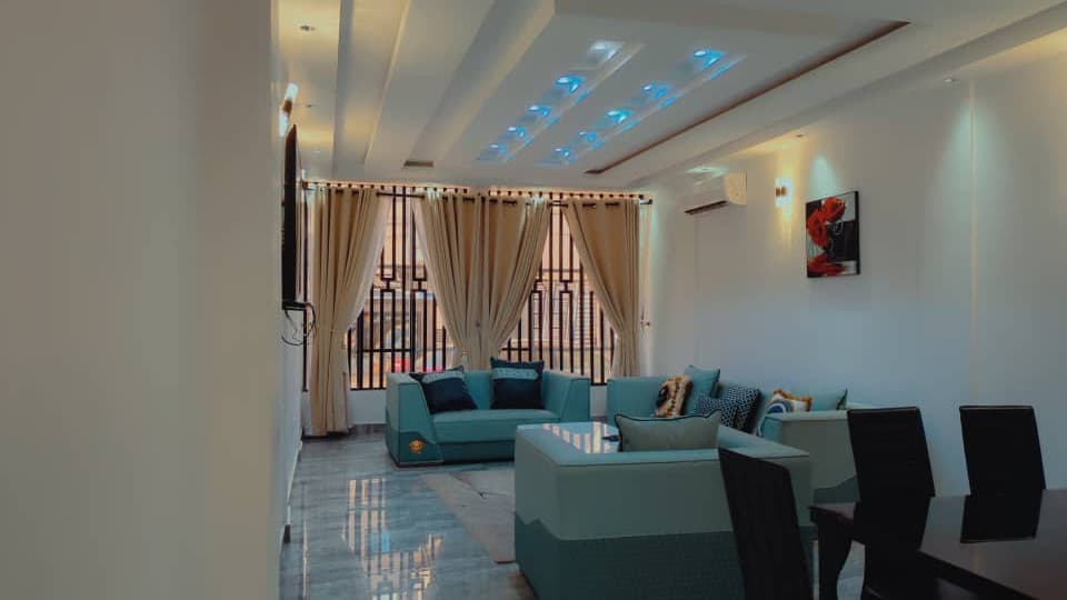 Salon de l’appartement A10 — canapés et fauteuils en velours bleu-vert, coussins baroques dorés et bleu nuit, sol en marbre, faux-plafond à gorge lumineuse bleue, climatiseur mural, doubles rideaux et coin repas au fond