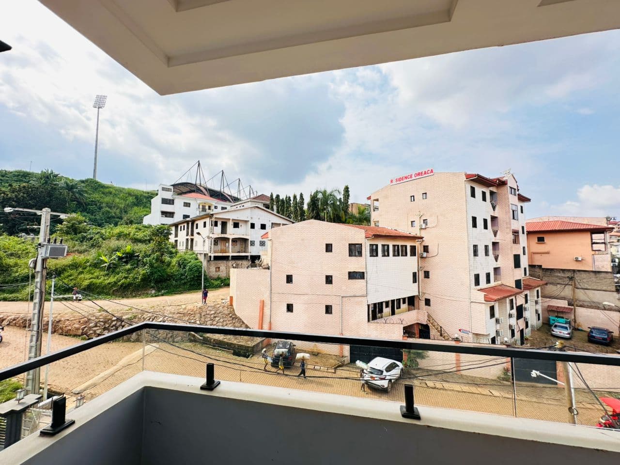 Vue depuis le balcon de l’appartement A40 — garde-corps en verre, route en contrebas, immeubles de quatre et cinq étages sur la colline d’en face, mât d’éclairage et toiture de stade sur la crête