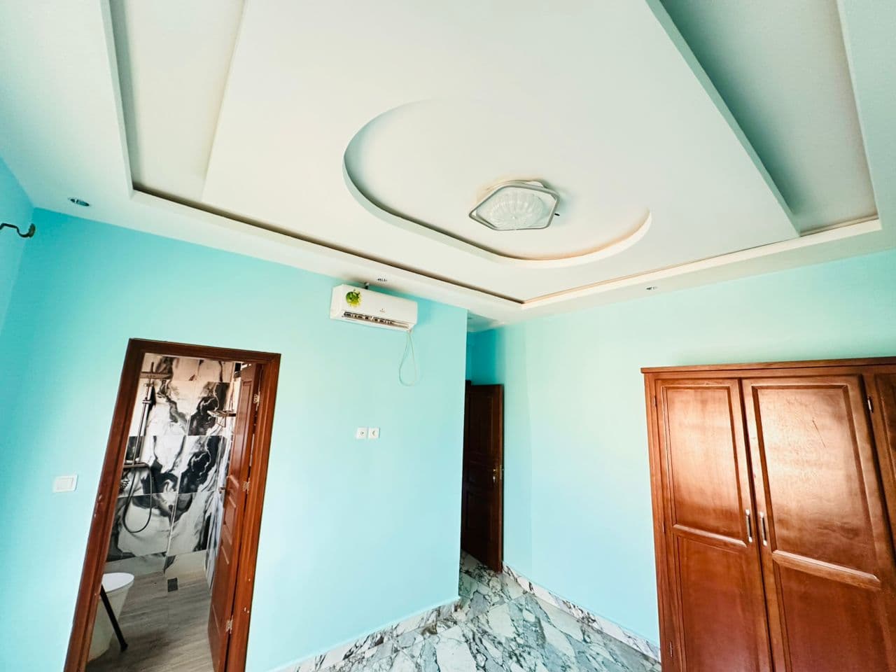 Chambre de l’appartement A40 aux murs turquoise — climatiseur, armoire double en bois verni, faux-plafond à voûte ovale et porte ouverte sur une salle de douche en marbre