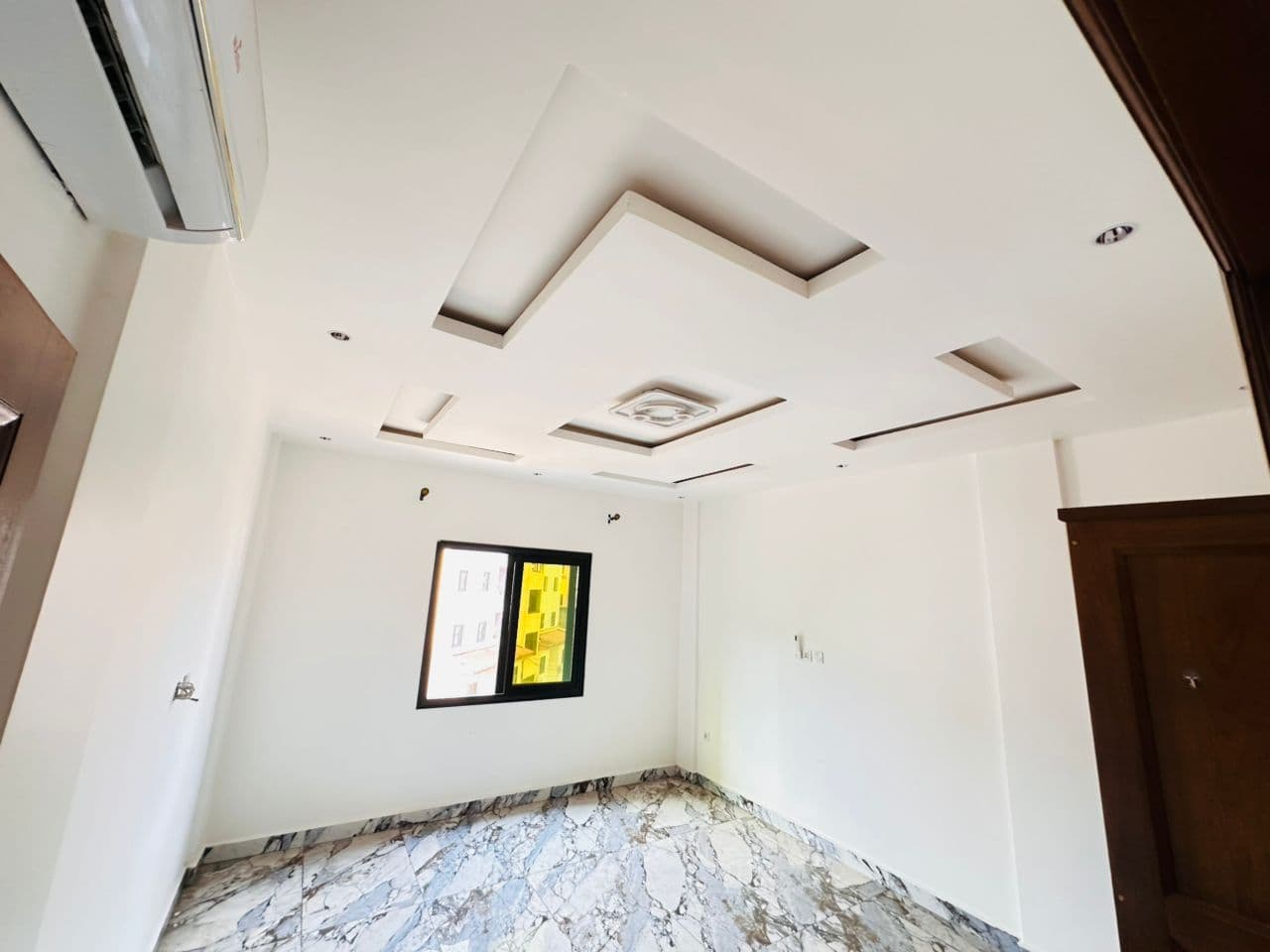 Chambre vide de l’appartement A40 aux murs blancs — fenêtre coulissante à châssis noir, faux-plafond à caisson central et spots encastrés, porte en bois verni et sol en marbre veiné