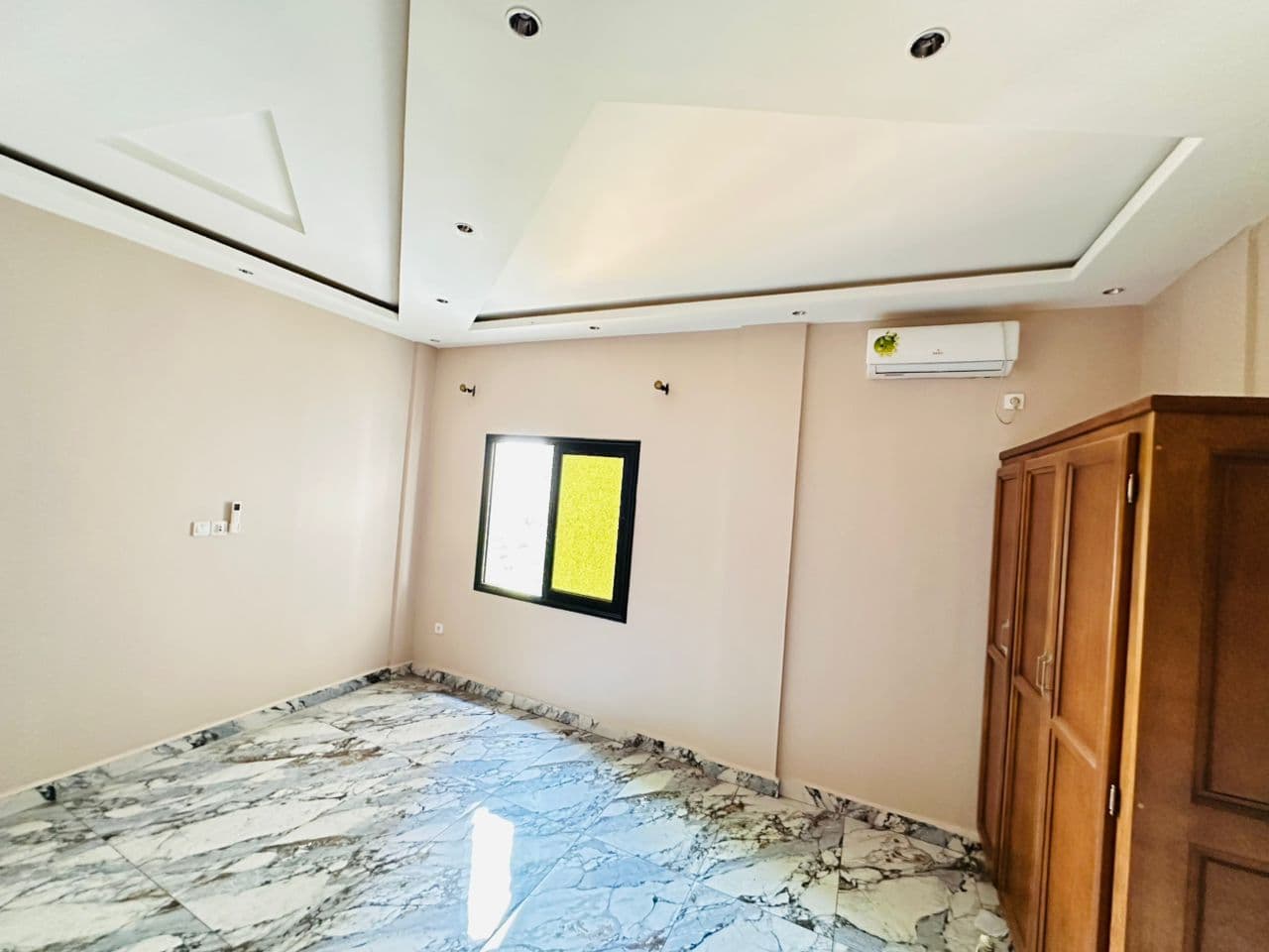 Chambre vide de l’appartement A40 aux murs taupe — climatiseur mural, fenêtre coulissante à châssis noir, grande armoire en bois verni, faux-plafond à pans et sol en marbre gris et blanc