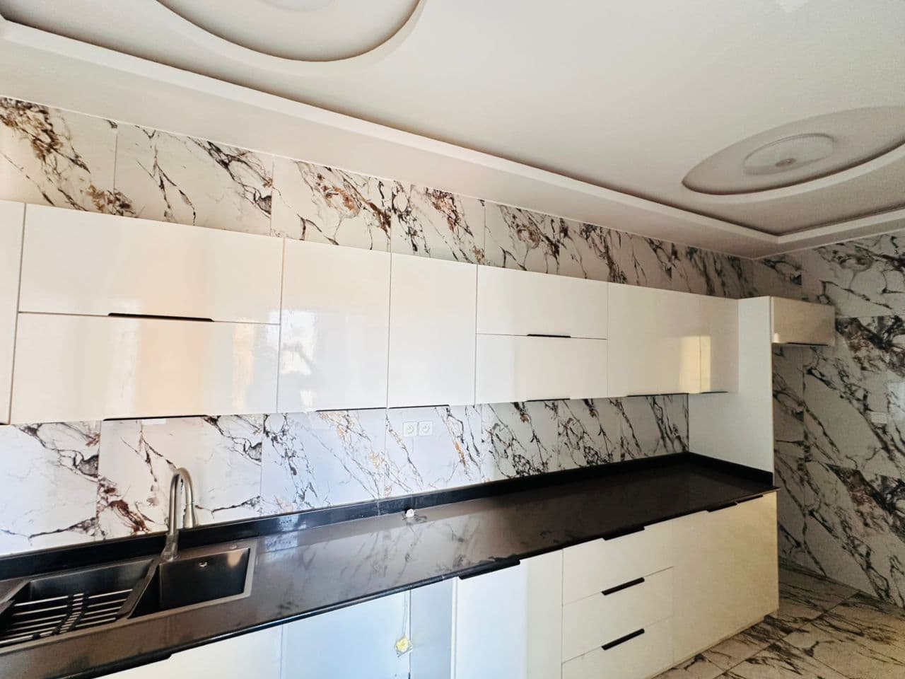 Cuisine de l’appartement A40 — meubles laqués blancs sans poignées en hauts et bas, crédence en marbre veiné toute hauteur, plan de travail en pierre noire, évier inox à égouttoir intégré et rosace lumineuse au plafond