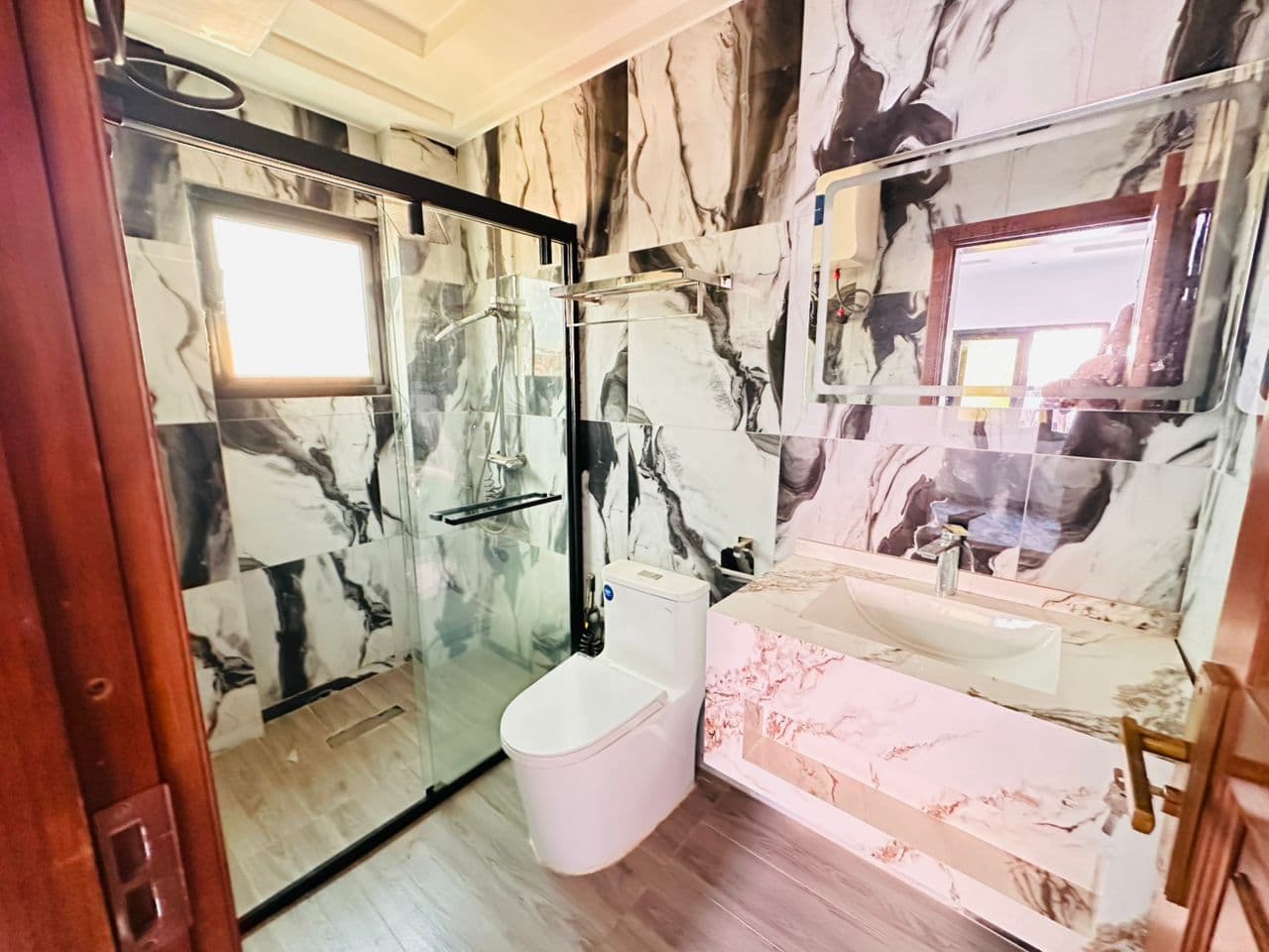Salle de douche de l’appartement A40 — marbre noir et blanc toute hauteur, cabine à paroi de verre coulissante avec ciel de pluie et douchette, WC, vasque à poser sur plan en marbre rosé et miroir rétroéclairé