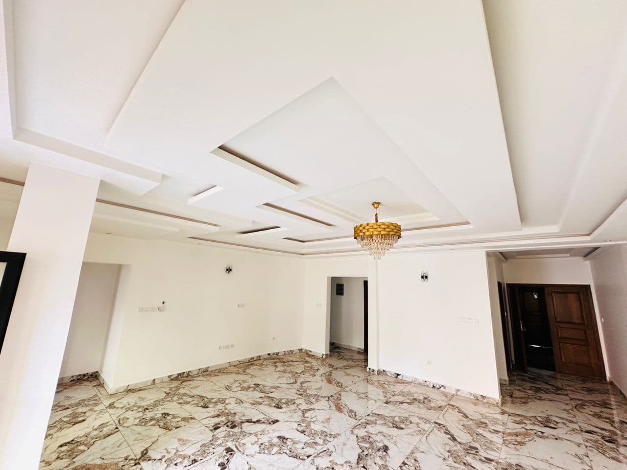 Salon de l’appartement A40 vu de l’autre extrémité — lustre doré à pampilles de cristal, faux-plafond à redans, poteau, porte en bois verni et sol en marbre veiné