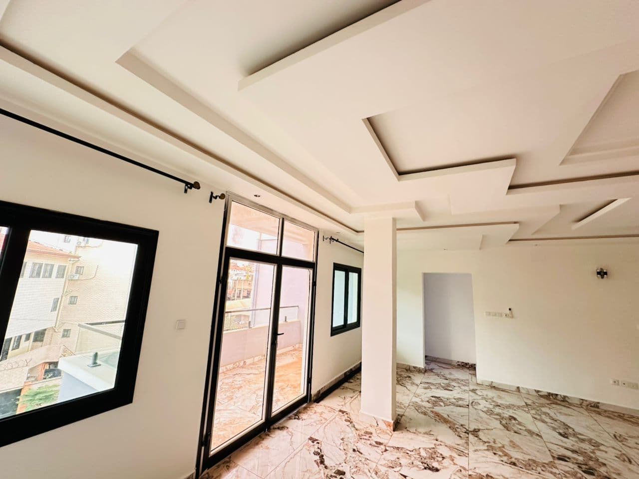 Salon vide de l’appartement A40 — sol en marbre veiné rose et or, faux-plafond à redans avec bandeaux lumineux, poteau central, baie vitrée à châssis noir ouvrant sur le balcon et tringles à rideaux posées