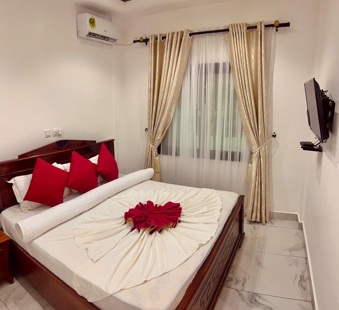 Première chambre du B35 — grand lit en bois verni, linge blanc et coussins rouges, serviette pliée en éventail, climatiseur mural, téléviseur mural et doubles rideaux dorés