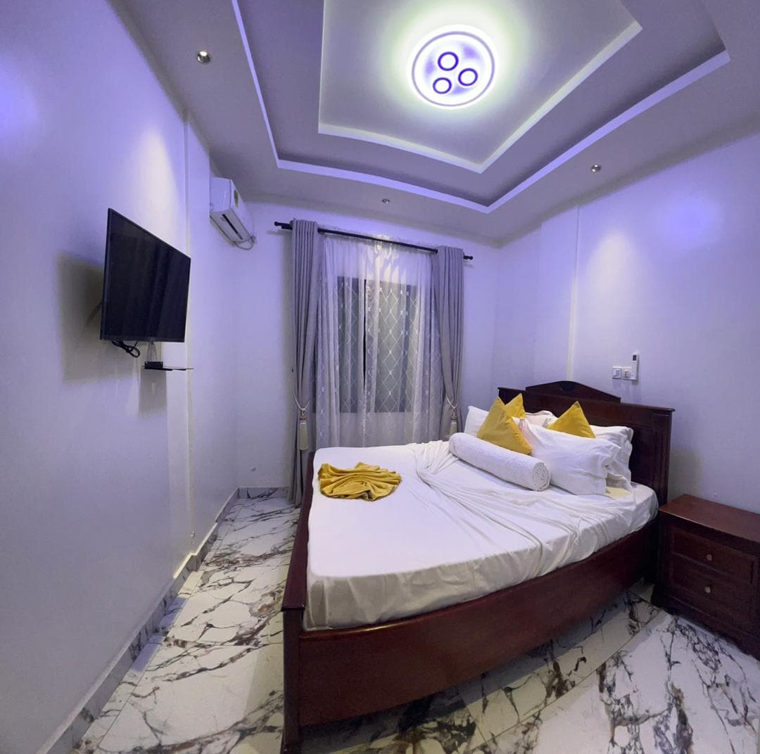 Seconde chambre du B35 — grand lit en bois verni, linge blanc et coussins jaunes, table de chevet, téléviseur mural, climatiseur, plafonnier circulaire à LED et sol en marbre veiné