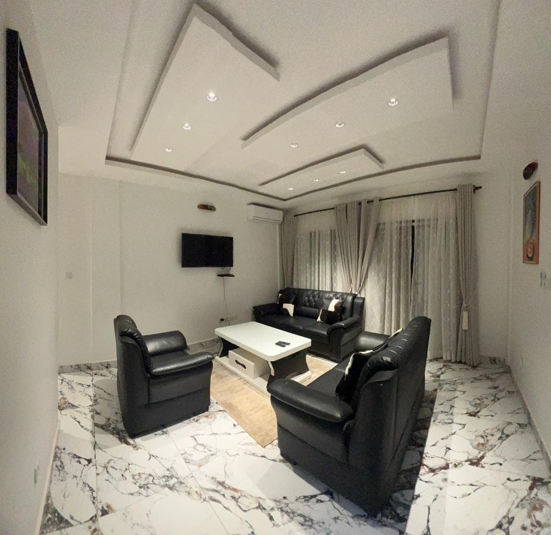 Salon du B35 — canapé et fauteuils en cuir noir capitonné, table basse blanche, téléviseur mural, climatiseur, faux-plafond à caissons et spots encastrés, sol en marbre blanc veiné