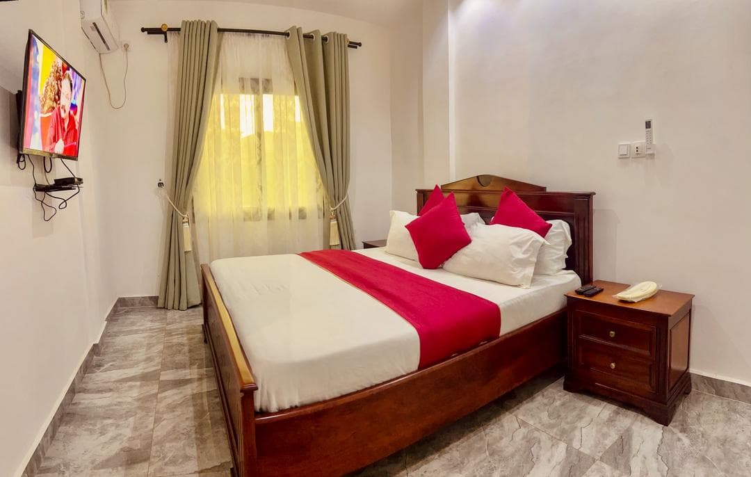 Chambre du studio SS140 — grand lit en bois verni, linge blanc et coussins framboise, table de chevet, téléviseur mural, climatiseur et rideaux voilés