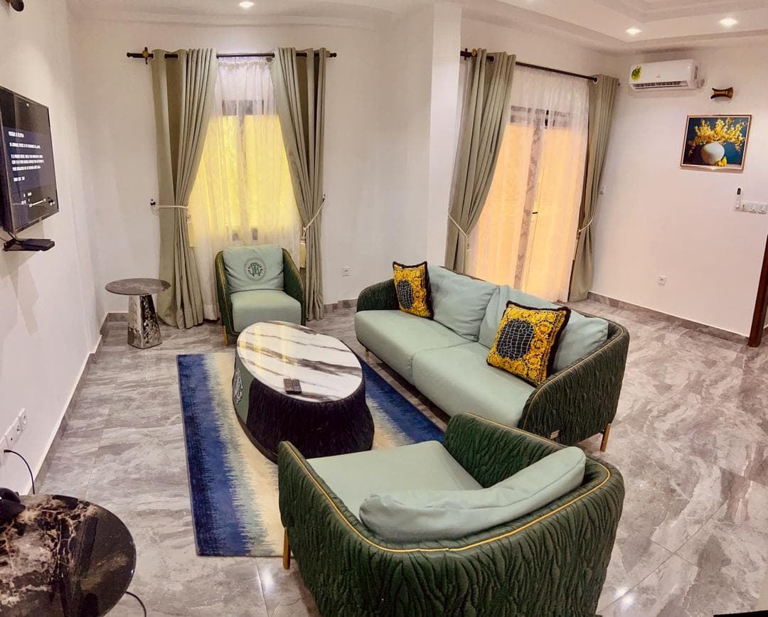 Salon du studio SS140 — canapé et fauteuils en velours vert d’eau, coussins baroques dorés, table basse ovale en marbre, sol en marbre gris, téléviseur mural et climatiseur