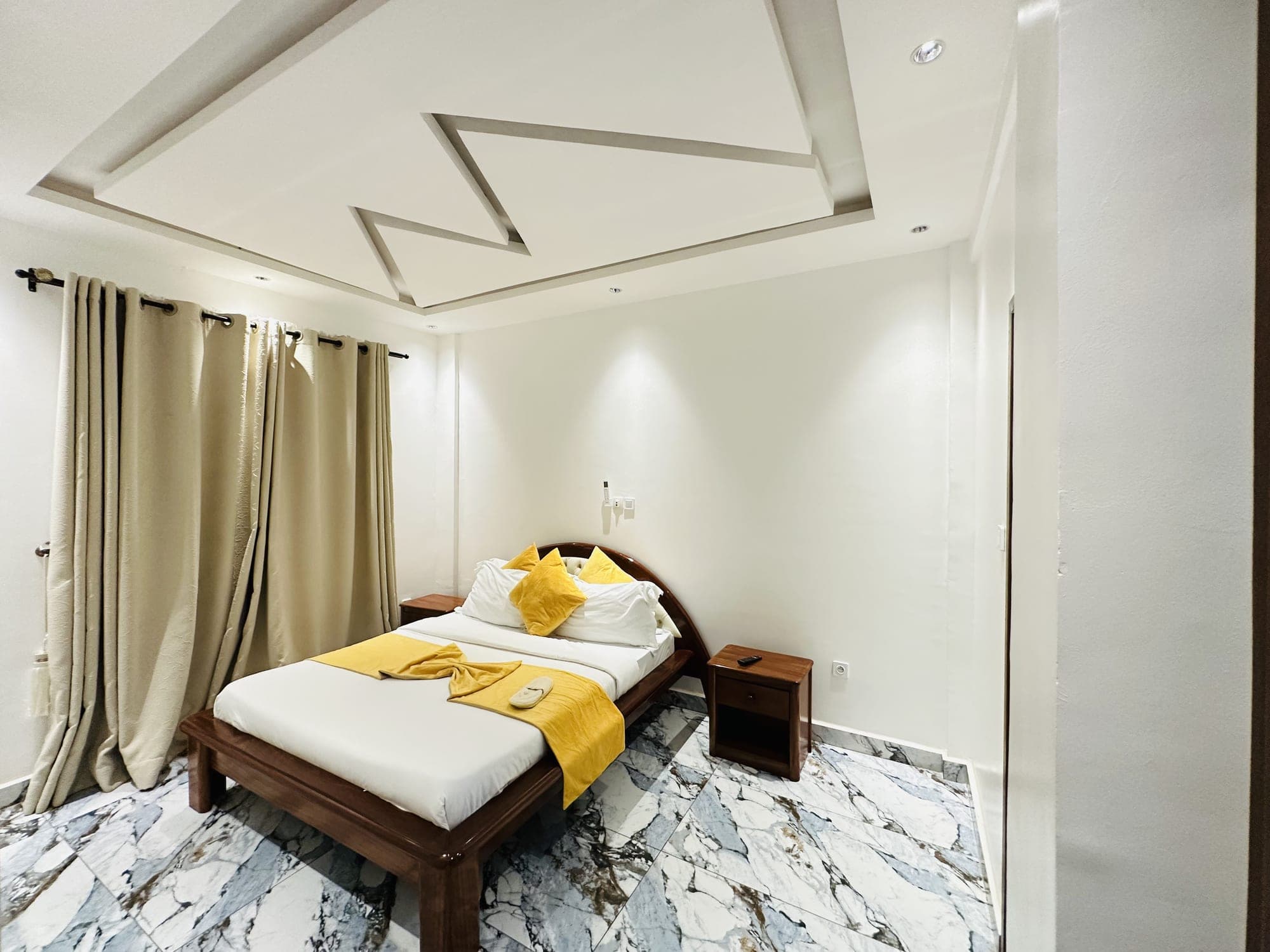 Chambre du studio ST005 — le grand lit dressé de blanc et de jaune moutarde entre deux tables de chevet en bois, rideaux beiges et sol en marbre veiné