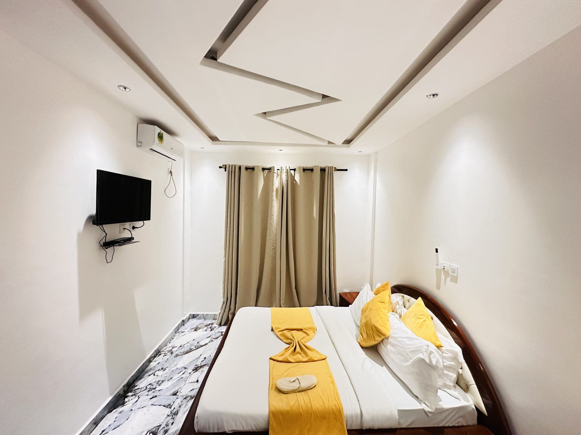 Chambre du studio ST005 — téléviseur mural et climatiseur face au lit, rideaux beiges tirés derrière la tête de lit, chaussons posés sur le jeté jaune