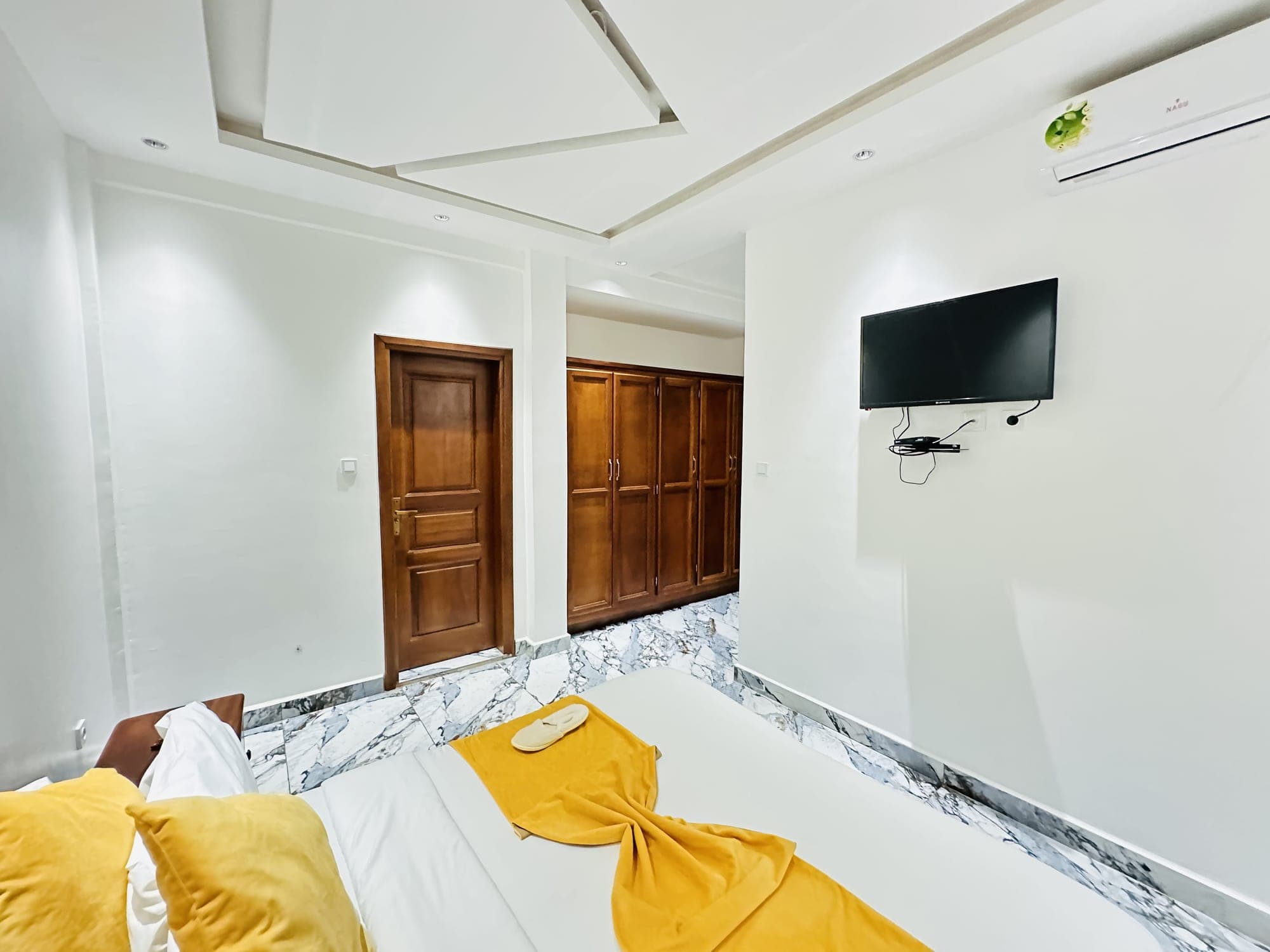 Rangements de la chambre du studio ST005 — armoire à quatre portes en bois verni, téléviseur mural, climatiseur et pied du lit avec serviette jaune pliée
