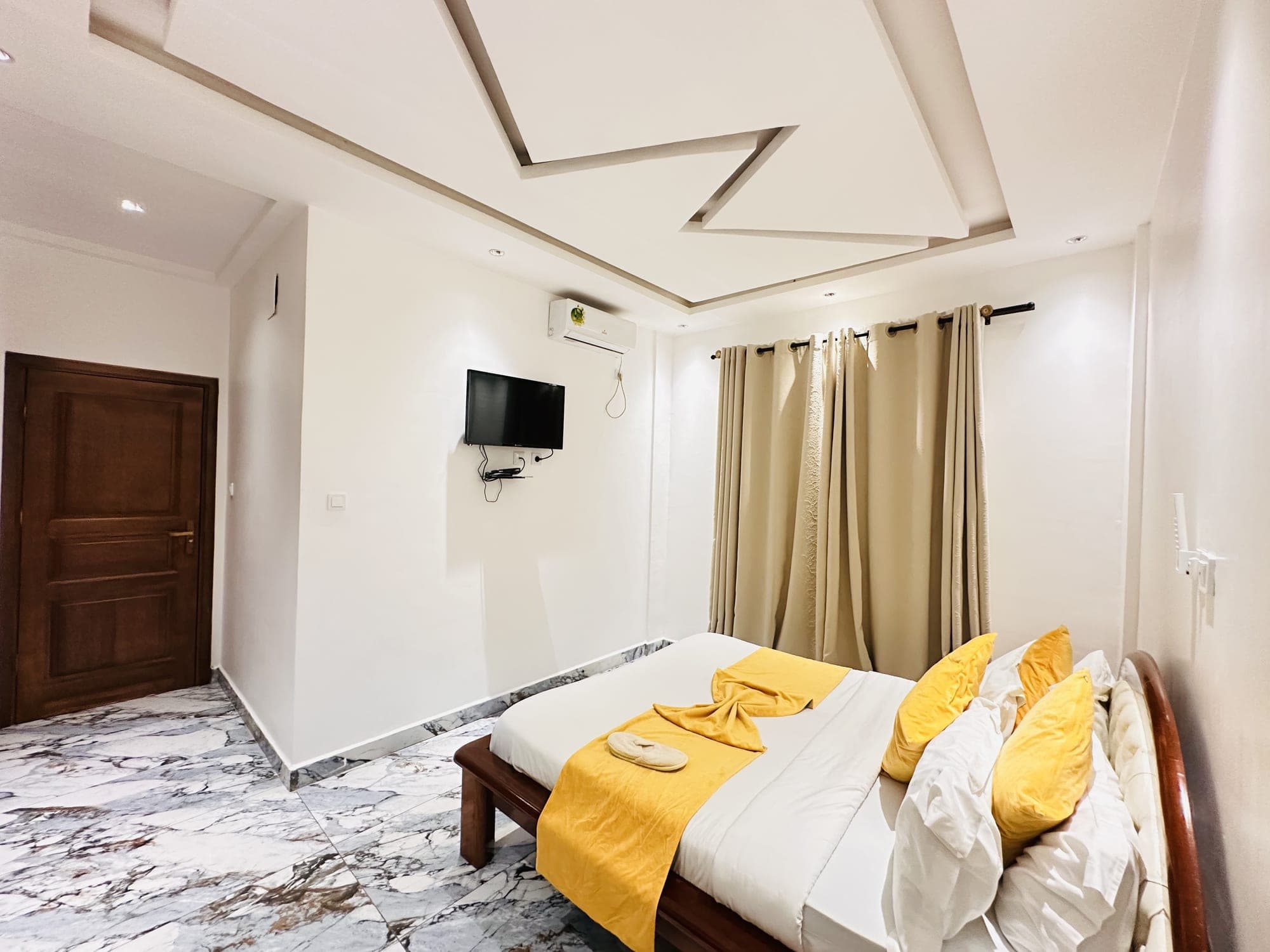 Chambre du studio ST005 — grand lit en bois verni, linge blanc et jeté jaune moutarde, téléviseur mural, climatiseur, doubles rideaux beiges et faux-plafond nervuré