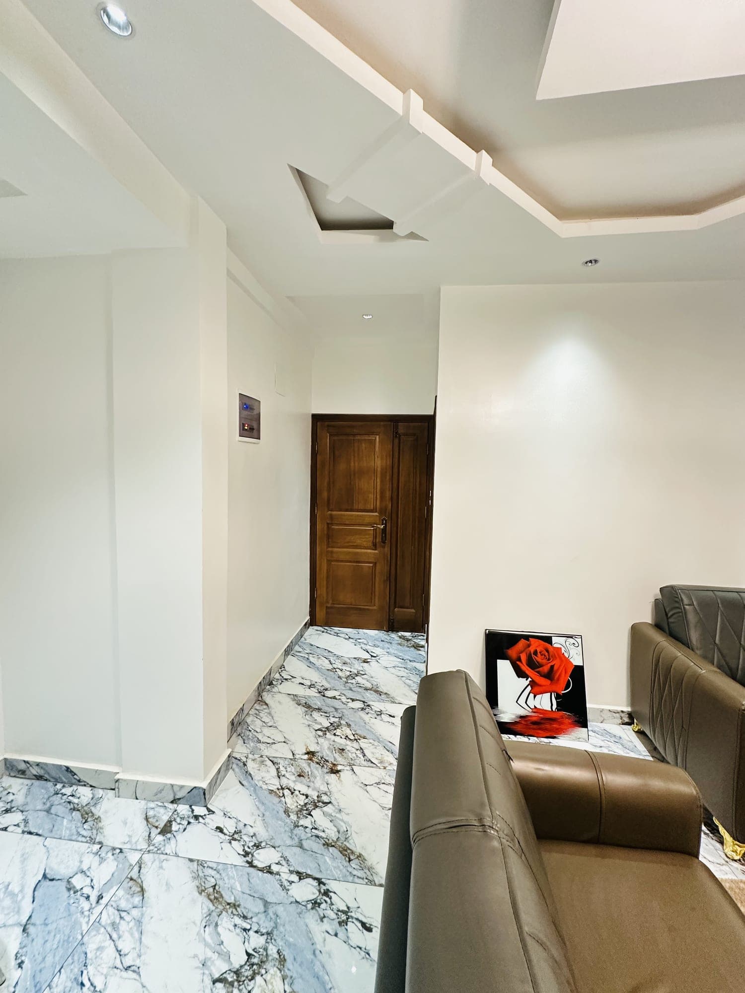 Entrée du studio ST005 — dégagement carrelé de marbre menant à la porte palière en bois verni, faux-plafond à caissons, spots encastrés et interphone mural
